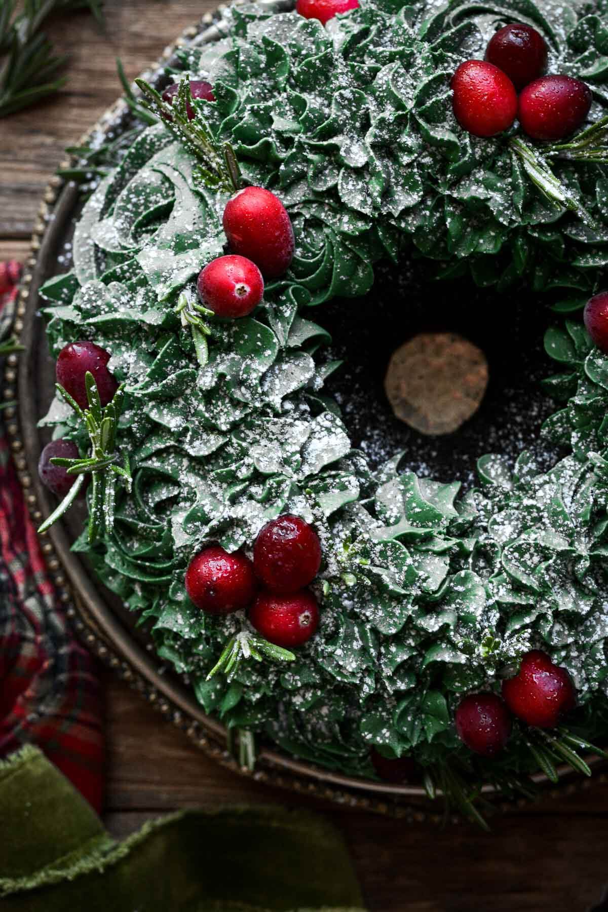Christmas wreath bundt cake decorated with piped green buttercream, cranberries and sprigs of rosemary.