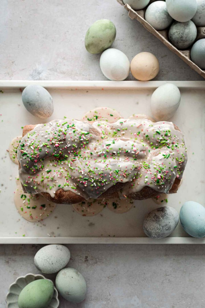 Loaf of Easter bread with icing and sprinkles, surrounded by dyed eggs.