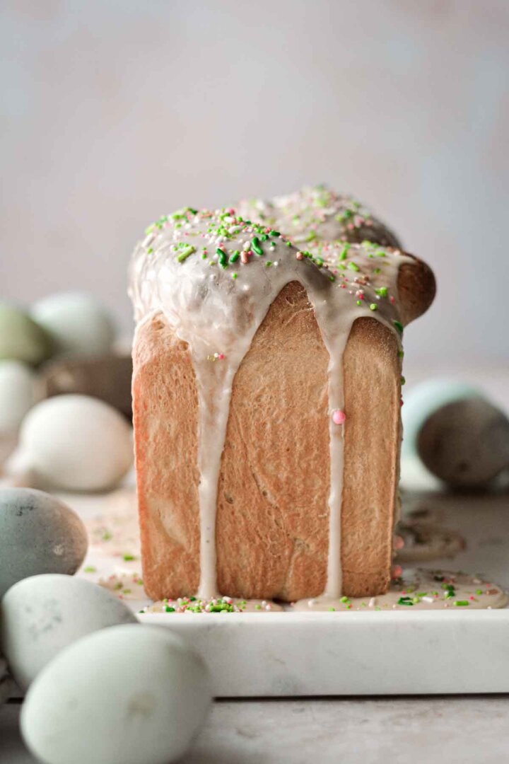 Loaf of Easter bread with icing and sprinkles, surrounded by dyed eggs.