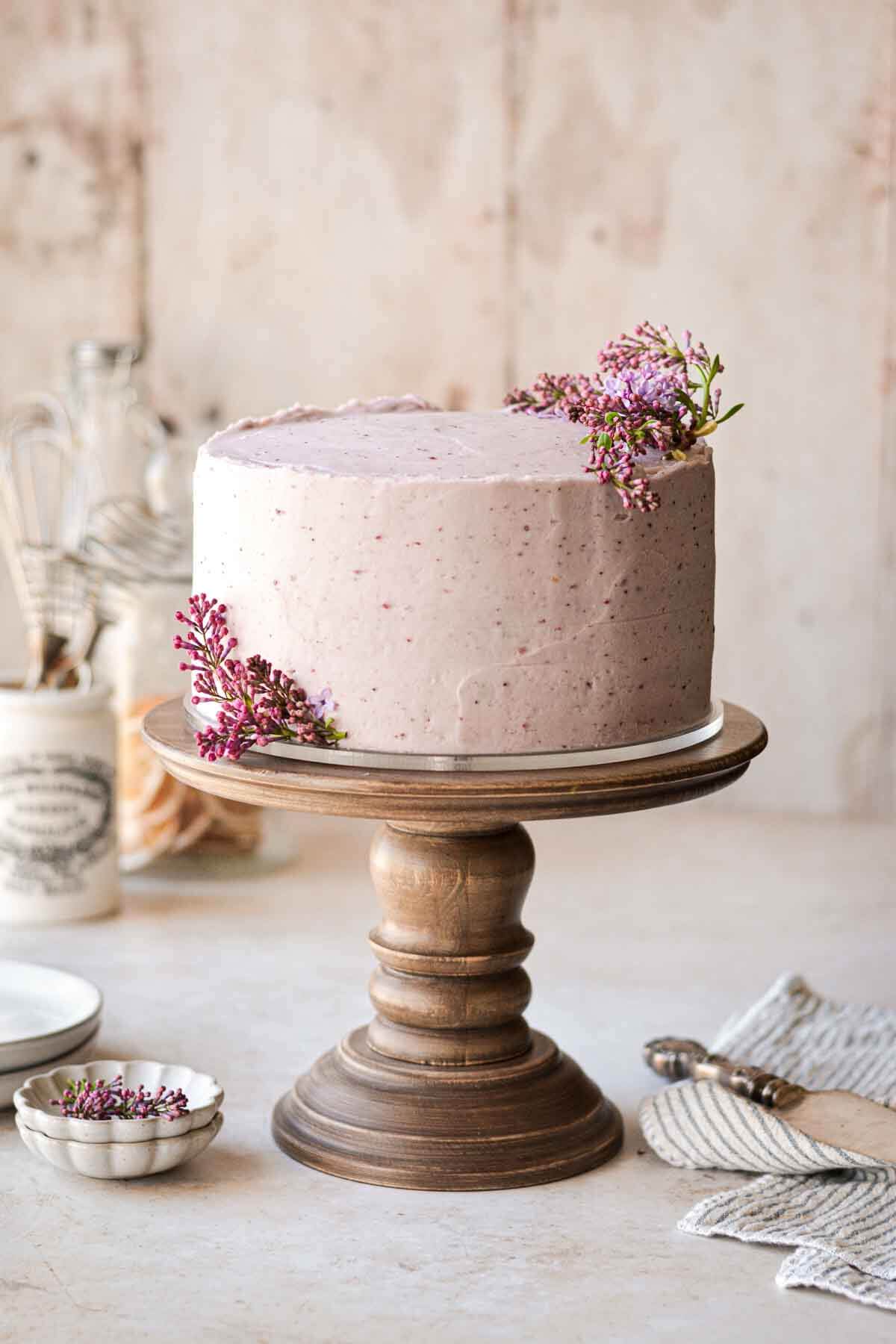 Fresh lilacs on a lemon blueberry layer cake, sitting on a wooden cake stand.