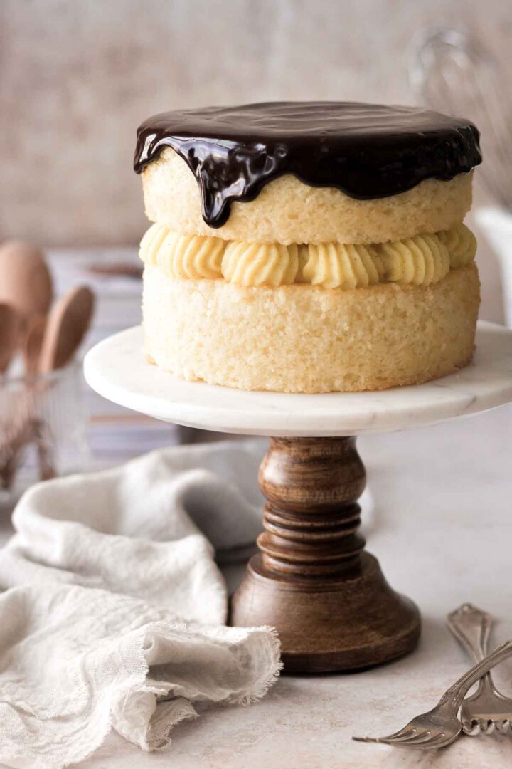 Boston cream pie with dripping ganache, on a marble and wood cake stand.