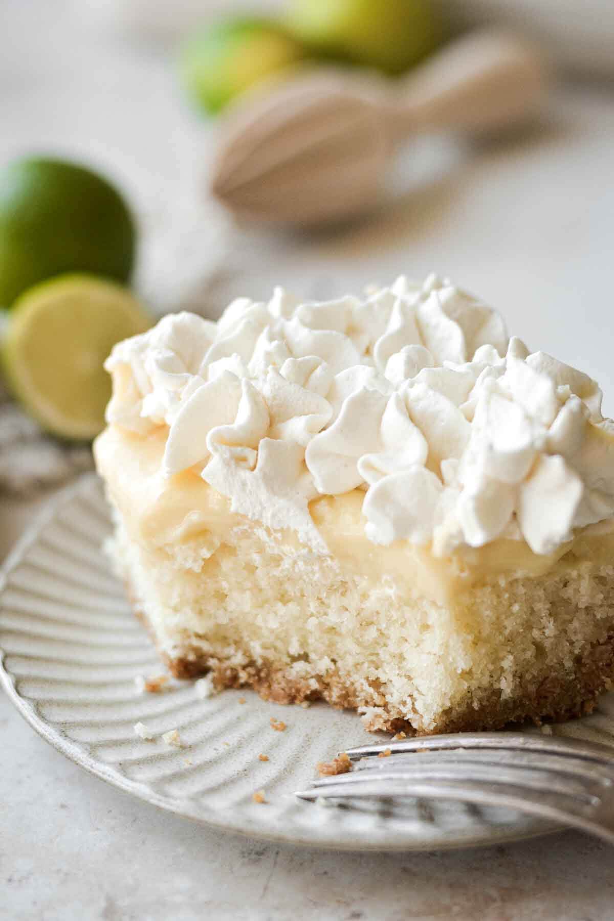 Slice of key lime pie cake, with layers of graham cracker crust, vanilla cake, lime pudding, and whipped cream.