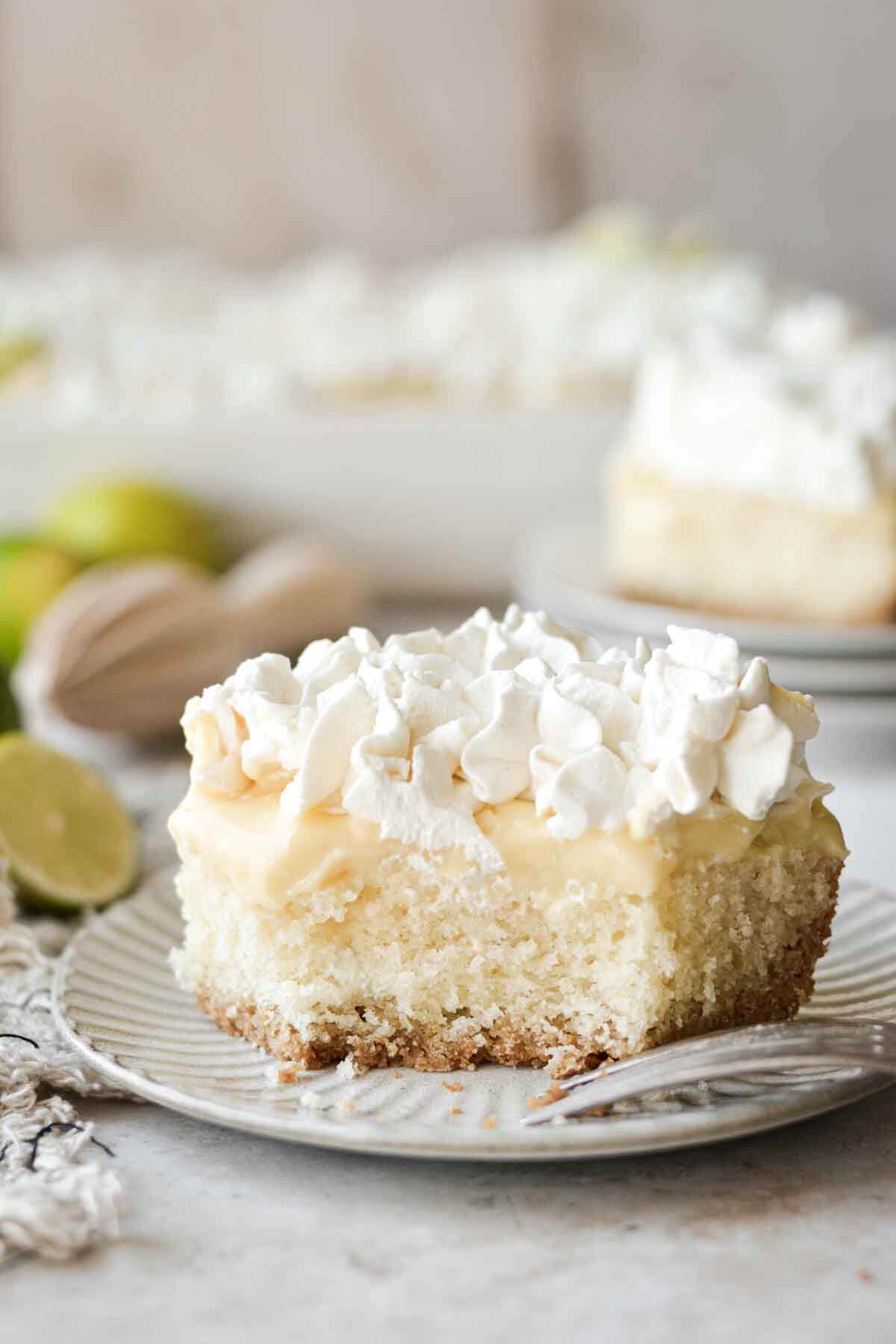 Slice of key lime pie cake, with layers of graham cracker crust, vanilla cake, lime pudding, and whipped cream.