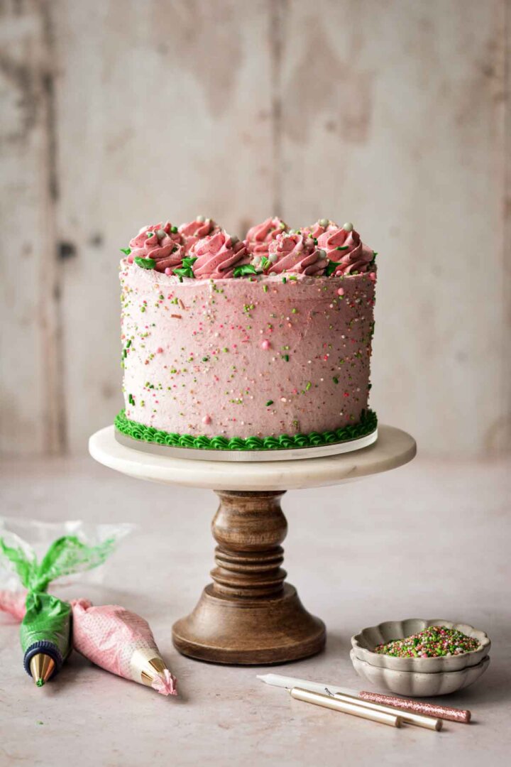 Strawberry cake, with pink and green buttercream and sprinkles, on a marble and wood cake stand.