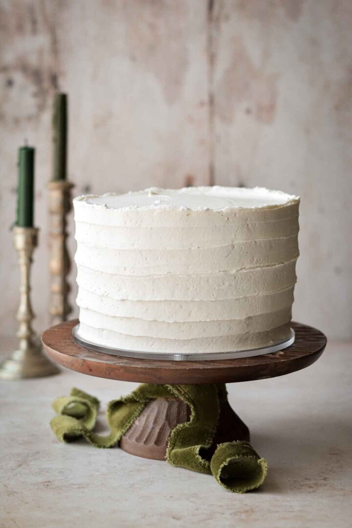 White velvet cake on a wooden cake stand, with green ribbon and candles.