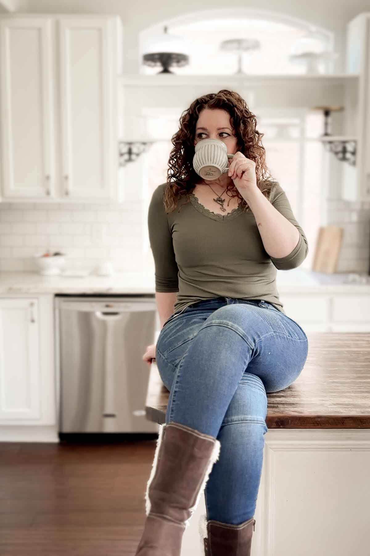 A woman sitting on a kitchen counter, sipping from a coffee cup.