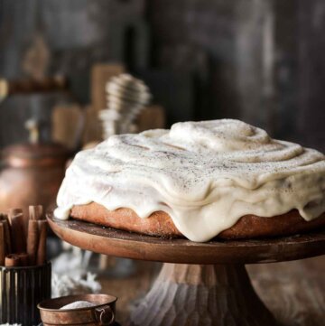 A giant cinnamon roll cake on a wooden cake stand with dripping cream cheese frosting.