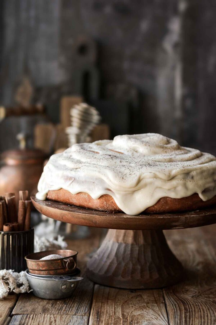 A giant cinnamon roll cake on a wooden cake stand with dripping cream cheese frosting.