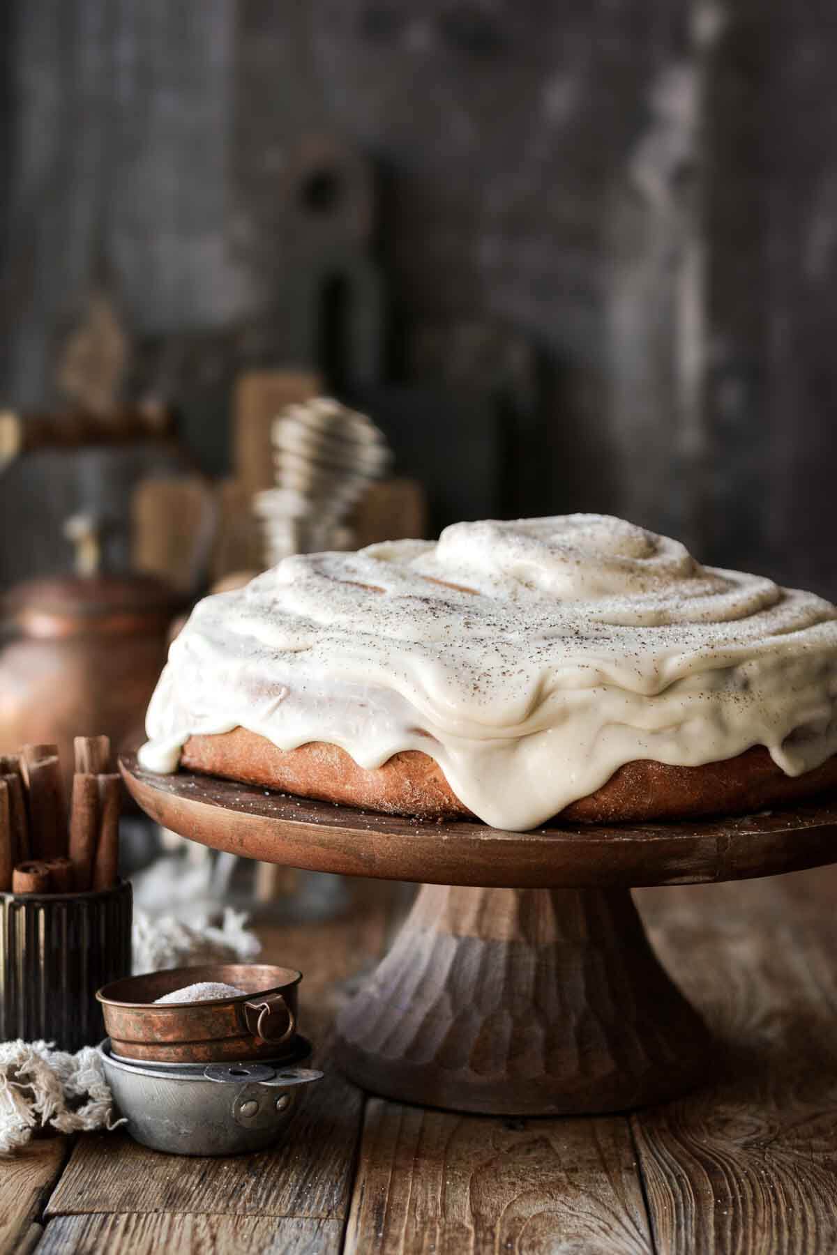 A giant cinnamon roll cake on a wooden cake stand with dripping cream cheese frosting.